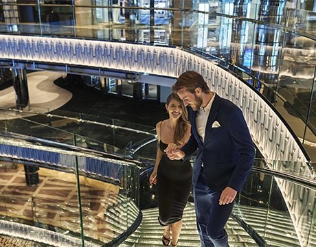 Elegantly dressed couple ascending a glass staircase aboard a luxurious cruise ship | MSC Cruises Elegantly dressed couple ascending a glass staircase aboard a luxurious cruise ship | MSC Cruises